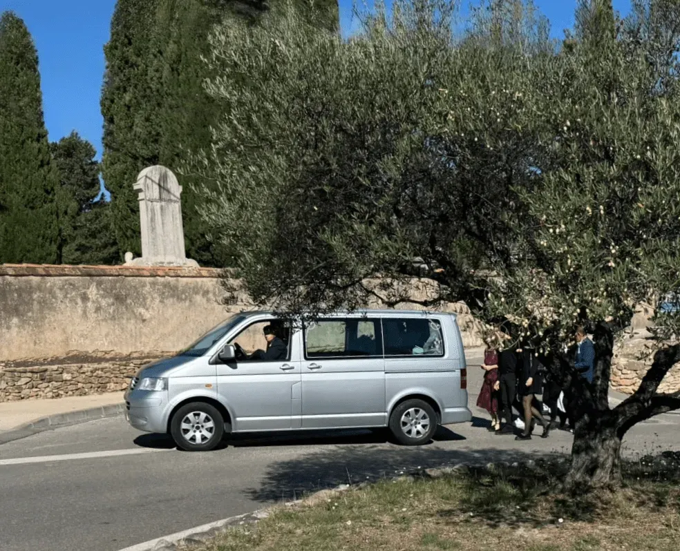 Fourgon funéraire gris roulant près d'un olivier lors d'un convoi d'obsèques.