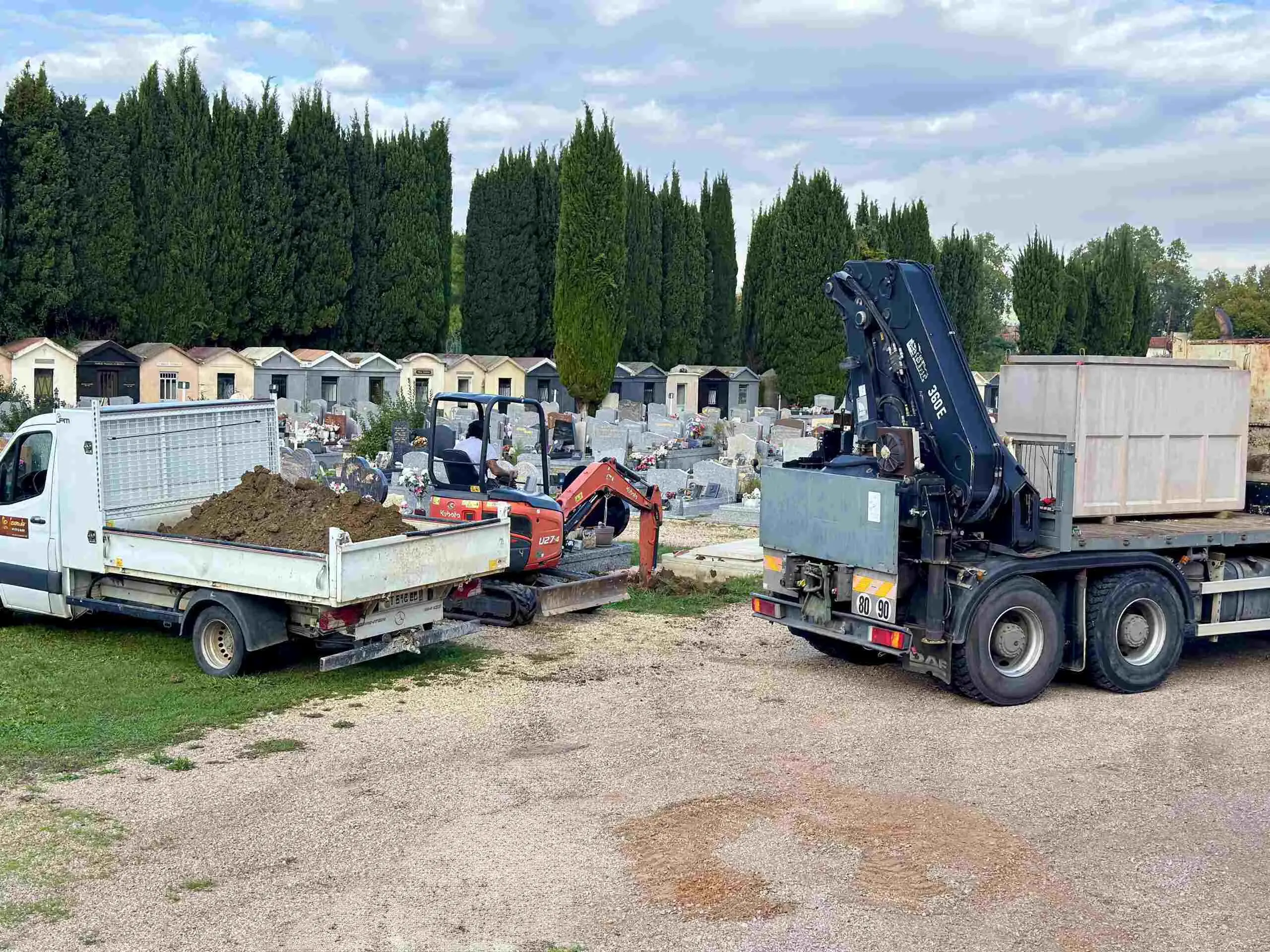 Camion et pelleteuse effectuant des travaux de marbrerie dans un cimetière.