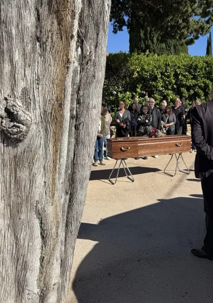 Cérémonie funéraire en extérieur autour d'un cercueil en bois placé sous des arbres.